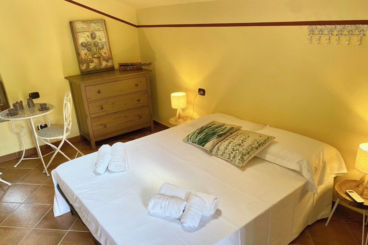Una cama bien hecha con sábanas blancas y dos toallas enrolladas, un aparador de madera con una pequeña mesa y sillas, y una lámpara montada en la pared.