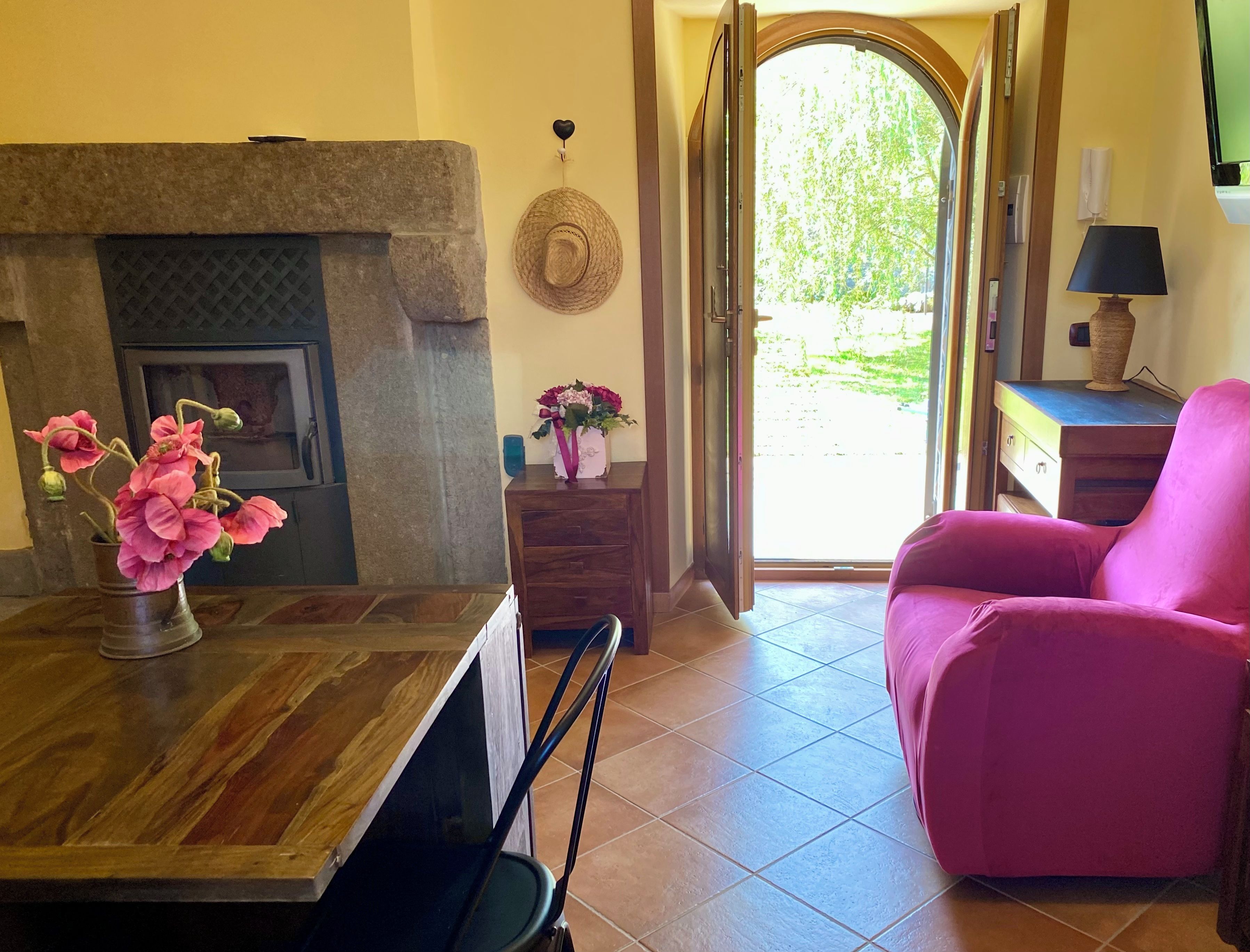 Una habitación con chimenea, un sillón rosa, una mesa de madera con un florero y vistas a través de una puerta abierta.
