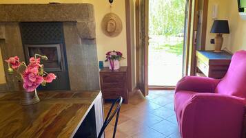 Una habitación con chimenea, un sillón rosa, una mesa de madera con un florero y vistas a través de una puerta abierta.