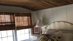 Quarto com cama, luminária de cabeceira, janela com persianas e tapete listrado colorido.