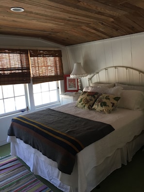 A bedroom with a bed, a bedside lamp, a window with blinds, and a colorful striped rug. - Artsy Lakeside Cottage w/ Deck & Woods Views — Pet Friendly, Sleeps 4 (Lakemont)