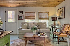 A cozy living room with a sofa, a coffee table with a bottle and glasses, a chair, and a lamp. - Artsy Lakeside Cottage w/ Deck & Woods Views — Pet Friendly, Sleeps 4 (Lakemont)