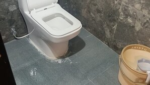 Eine weiße Toilette mit Tank und Sitz, ein grauer Fliesenboden und ein beiges Eimer mit der Aufschrift 'ARISTO', einem weißen Deckel und einem weißen Gegenstand darin.