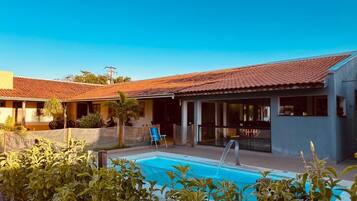 A pool area with a clear blue sky, a covered patio, and a building with a tiled roof.