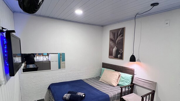 Quarto com cama, escrivaninha, cadeira, ventilador e TV fixada na parede.