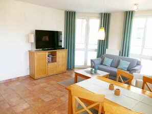 A living room with a television on a wooden cabinet, a grey sofa, a dining table with chairs, and a window with curtains. - Apartment Building Linquenda 12 (Heringsdorf)