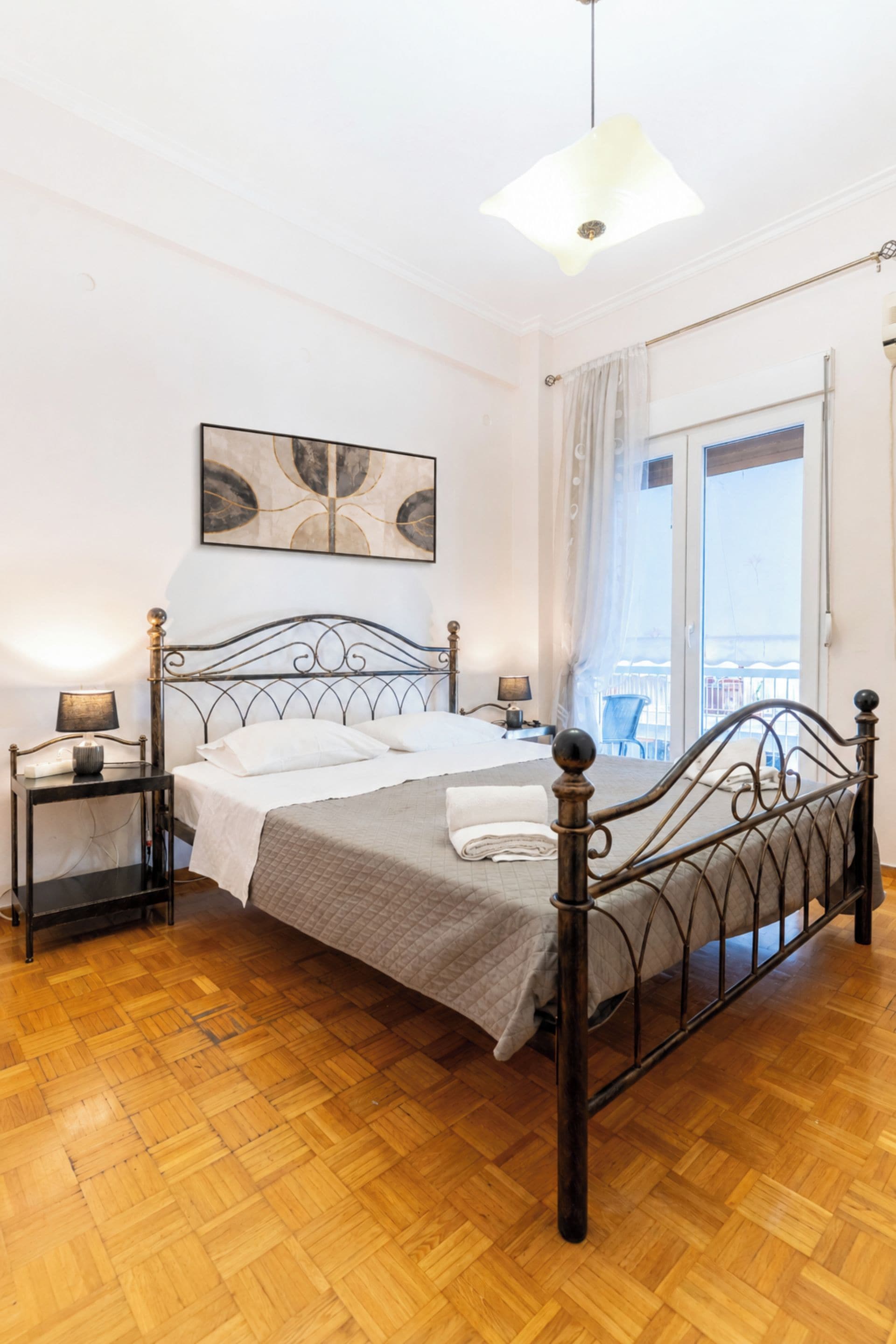 Un dormitorio con cama, mesitas de noche, lámpara, luz de techo, ventana con cortinas y una obra de arte colgada en la pared.