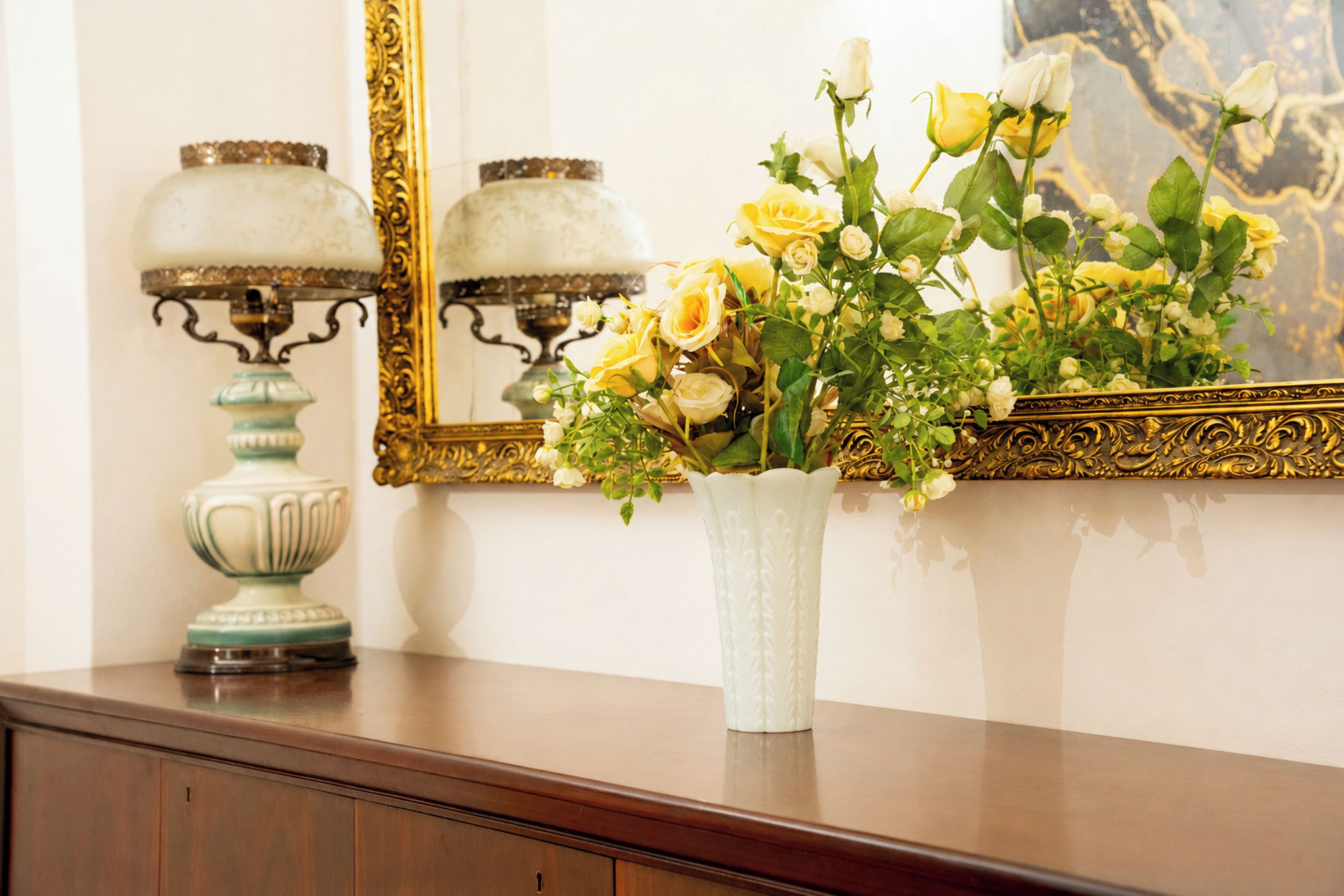 Un jarrón blanco con rosas amarillas sobre una superficie de madera, un espejo con marco dorado y dos lámparas de mesa ornamentadas.