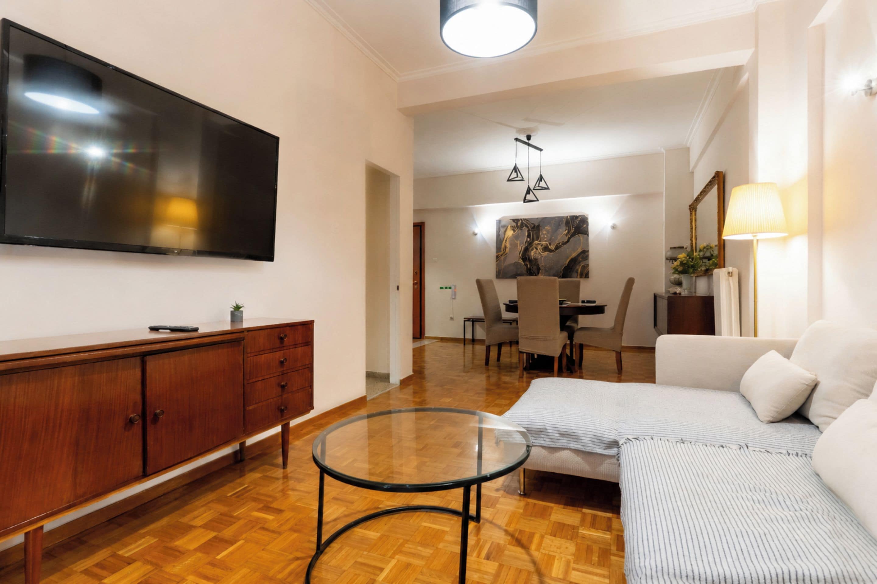 A living room with a flat-screen TV, a wooden cabinet, a glass coffee table, a sofa, and a dining area with chairs and a painting.