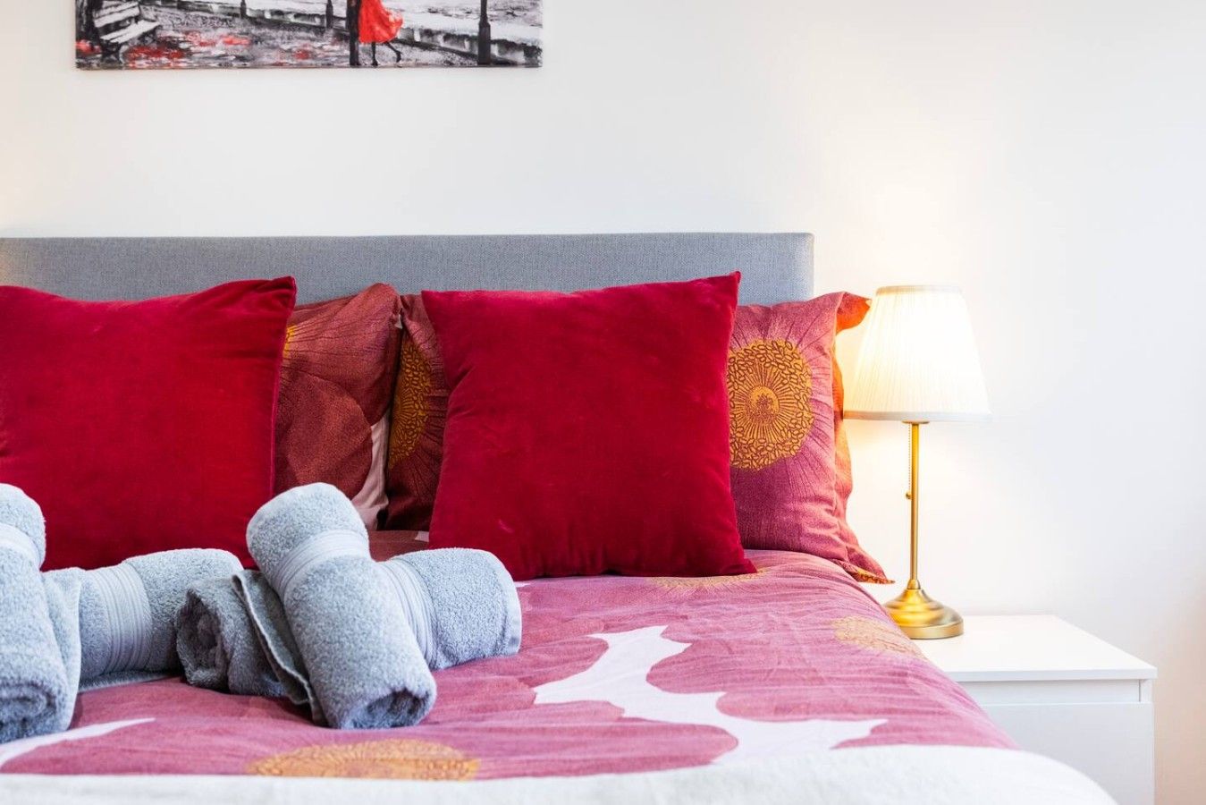 Un lit bien fait, agrémenté d’oreillers rouges et dorés, une table de chevet blanche avec une lampe, et un tableau accroché au mur.