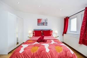 A bedroom with a large bed, two bedside tables with lamps, a window with red curtains, and a wall-mounted picture. - West Kensington 2BR | Sleeps 7 | Steps to Tube (Greater London)