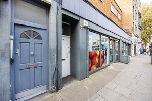 A building with a blue door, a window with a fruit advertisement, and a visible street. - West Kensington 2BR | Sleeps 7 | Steps to Tube (Greater London)