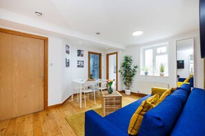 A room with a wooden floor, a blue sofa, a dining table with chairs, a potted plant, and a door. - West Kensington 2BR | Sleeps 7 | Steps to Tube (Greater London)