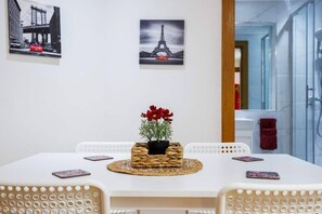 A dining area with a white table, four chairs, a woven placemat, and a potted plant with red flowers. - West Kensington 2BR | Sleeps 7 | Steps to Tube (Greater London)
