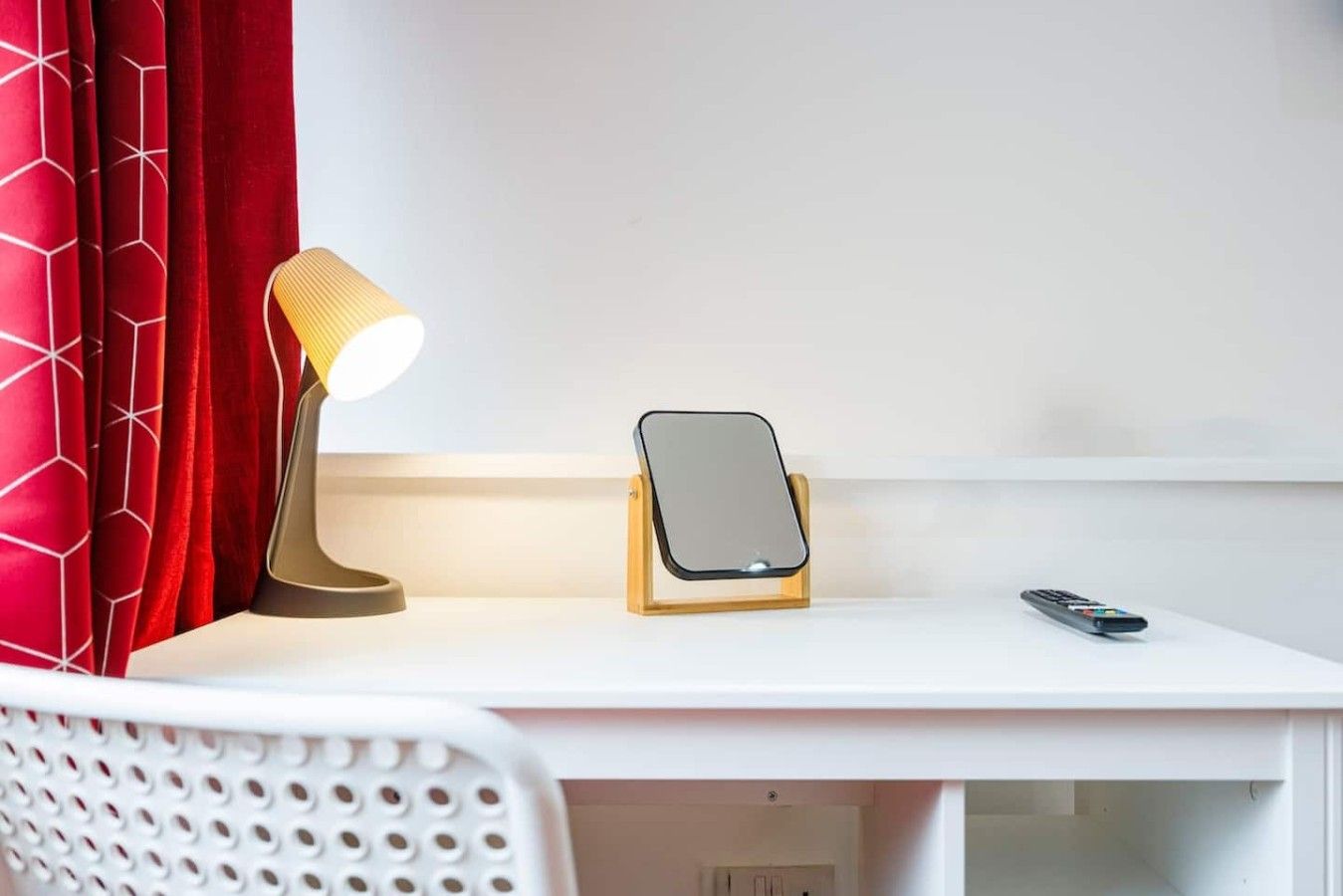 A white desk with a lamp, a smartphone on a stand, and a remote control.