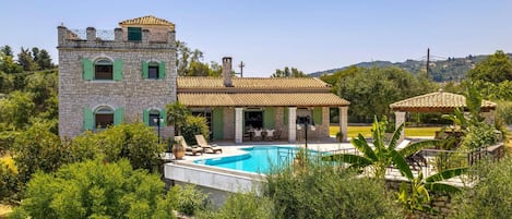A villa with a swimming pool, surrounded by lush greenery and a mountainous backdrop.
