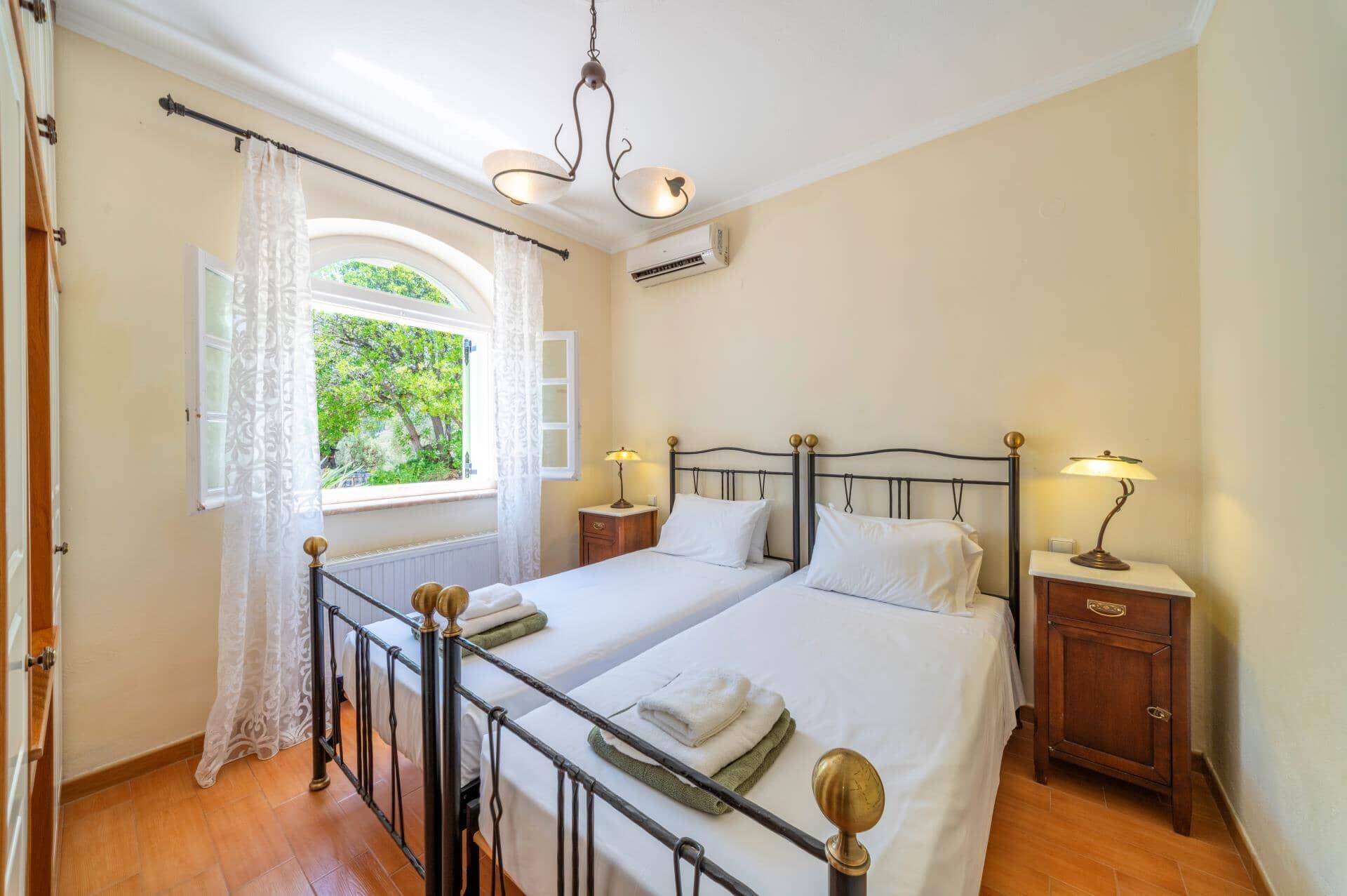 Una cama doble con estructura metálica, dos mesitas de noche con lámparas, un ventanal con cortinas y un equipo de aire acondicionado.
