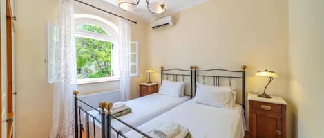 Een tweepersoonsbed met een metalen frame, twee nachtkastjes met lampen, een raam met gordijnen en een airconditioning.