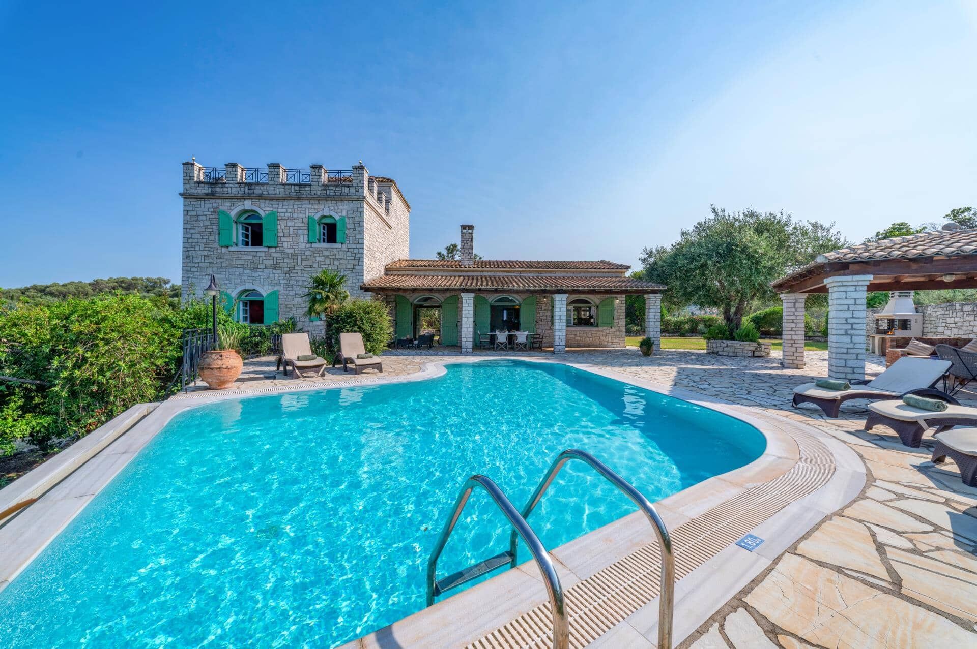 A swimming pool with a stone deck, a stone building with arched windows, and outdoor furniture.