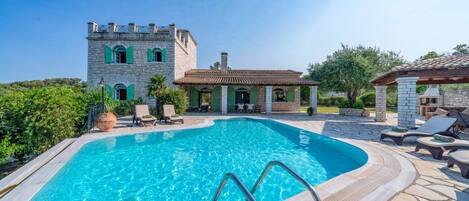 A swimming pool with a stone deck, a stone building with arched windows, and outdoor furniture.