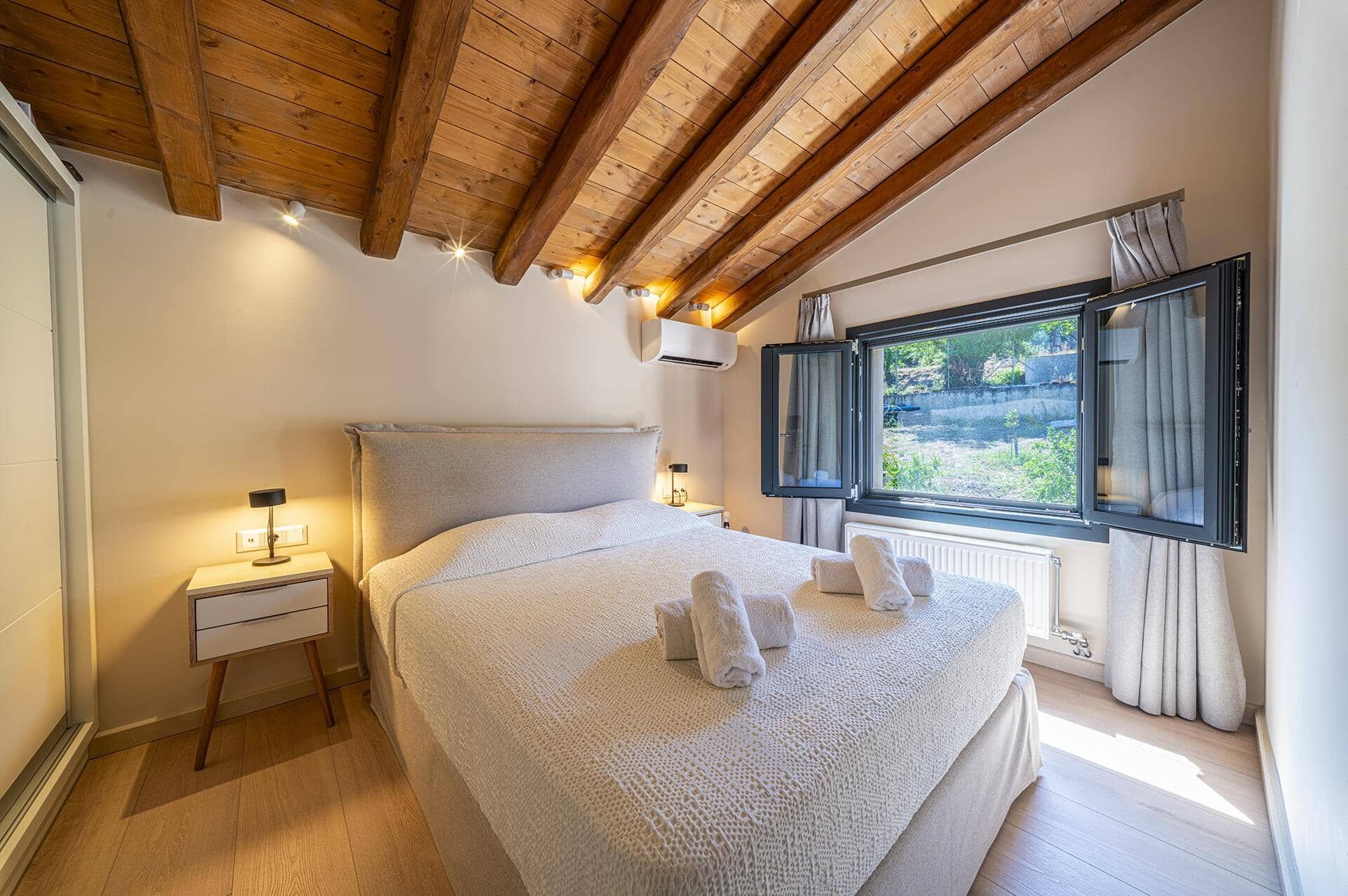Een slaapkamer met een houten plafond, een groot bed met witte beddengoed, een nachtkastje, een raam met gordijnen en een airconditioner.