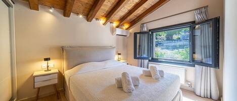 Un dormitorio con techo de madera, una cama grande con ropa blanca, una mesita de noche, una ventana con cortinas y un equipo de aire acondicionado.