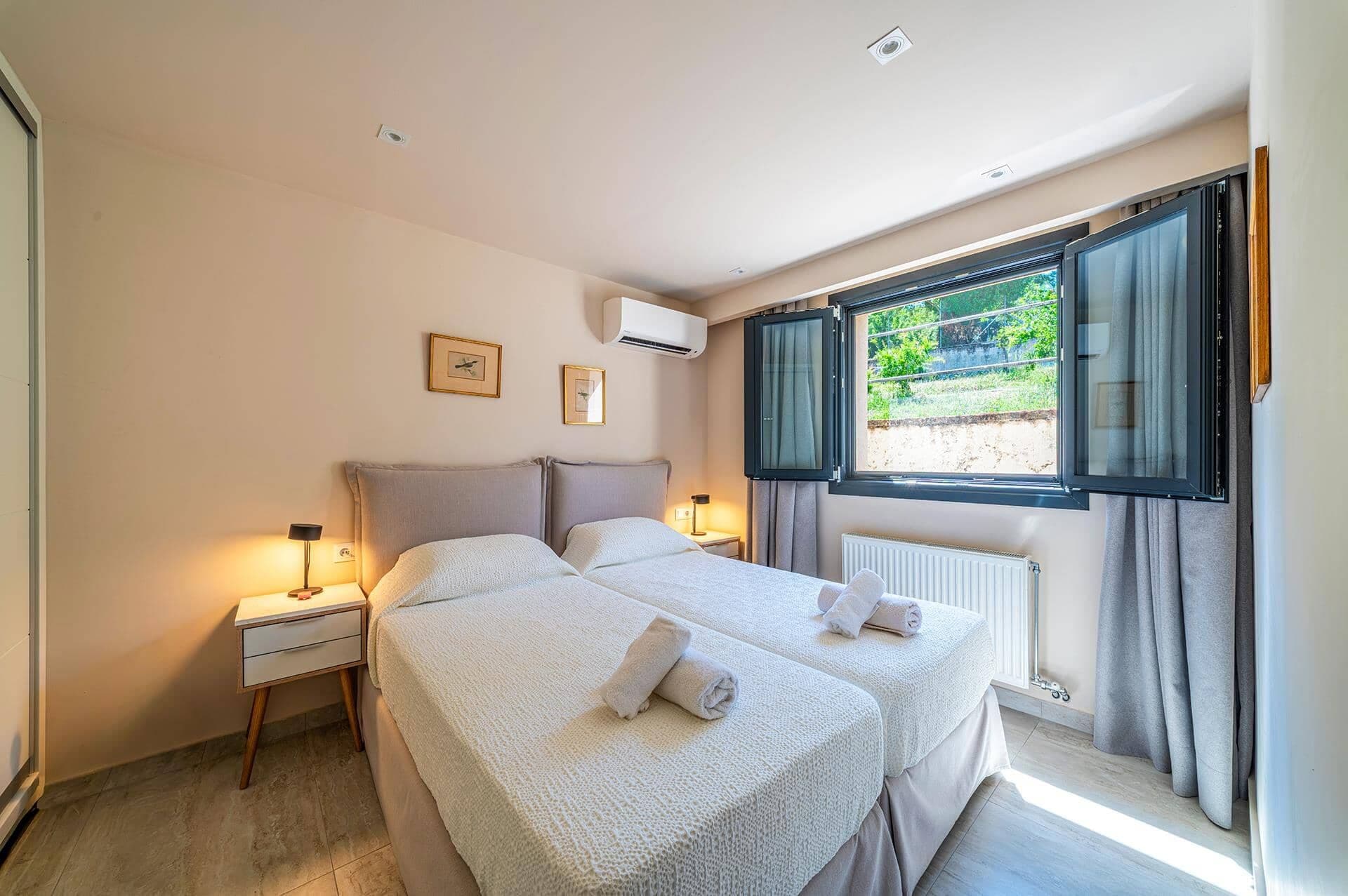 A bedroom with two beds, a window with curtains, an air conditioning unit, and a bedside table with a lamp.