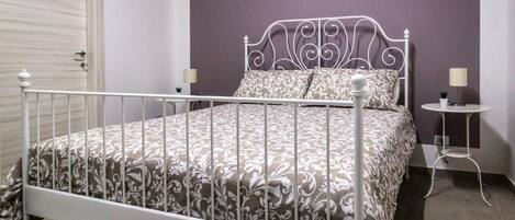 Ein Schlafzimmer mit einem Metallbettgestell, gemusterten Betttextilien, einem Nachttisch mit Lampe und einem an der Wand befestigten Regal.