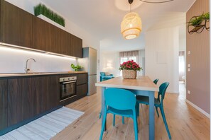A modern kitchen with a built-in oven, a dining area with a wooden table and teal chairs, and a hanging light fixture. - Cilea Apartment near Venice (Noale)
