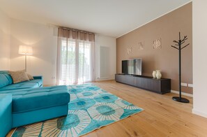 A modern living room with a blue sofa, a floor lamp, a flat-screen TV on a dark wooden cabinet, and a patterned rug. - Cilea Apartment near Venice (Noale)