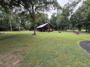 A rustic wooden cabin with a metal roof, surrounded by a well-maintained grassy area and trees. - Just West of Little Rock Retreat (Paron)