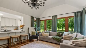 A spacious living area with a sofa, a dining table with bar stools, and a view of a green park through large windows.
