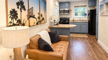 A modern living room with a leather sofa, a coffee table, and a lamp. There is a kitchen in the background with a microwave, stove, and cabinets.