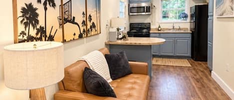 Un salon moderne avec un canapé en cuir, une table basse et une lampe. On aperçoit une cuisine en arrière-plan, avec un four à micro-ondes, une cuisinière et des placards.