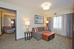 A hotel room with a sofa, ottoman, two side tables, a lamp, a framed picture, and a large window with curtains.
