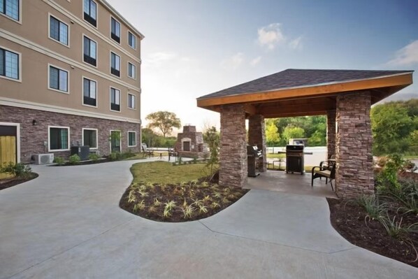 A covered outdoor seating area with a stone structure, a paved walkway, and a building with multiple windows in the background. - Accessible 2BR Suite | Free Daily Breakfast & Pool (Austin)