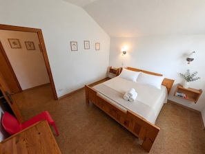 Ein Schlafzimmer mit einem hölzernen Bett, weißen Bettwäsche, einem Nachttisch mit Lampe sowie einem kleinen Regal mit Büchern und einer Pflanze.