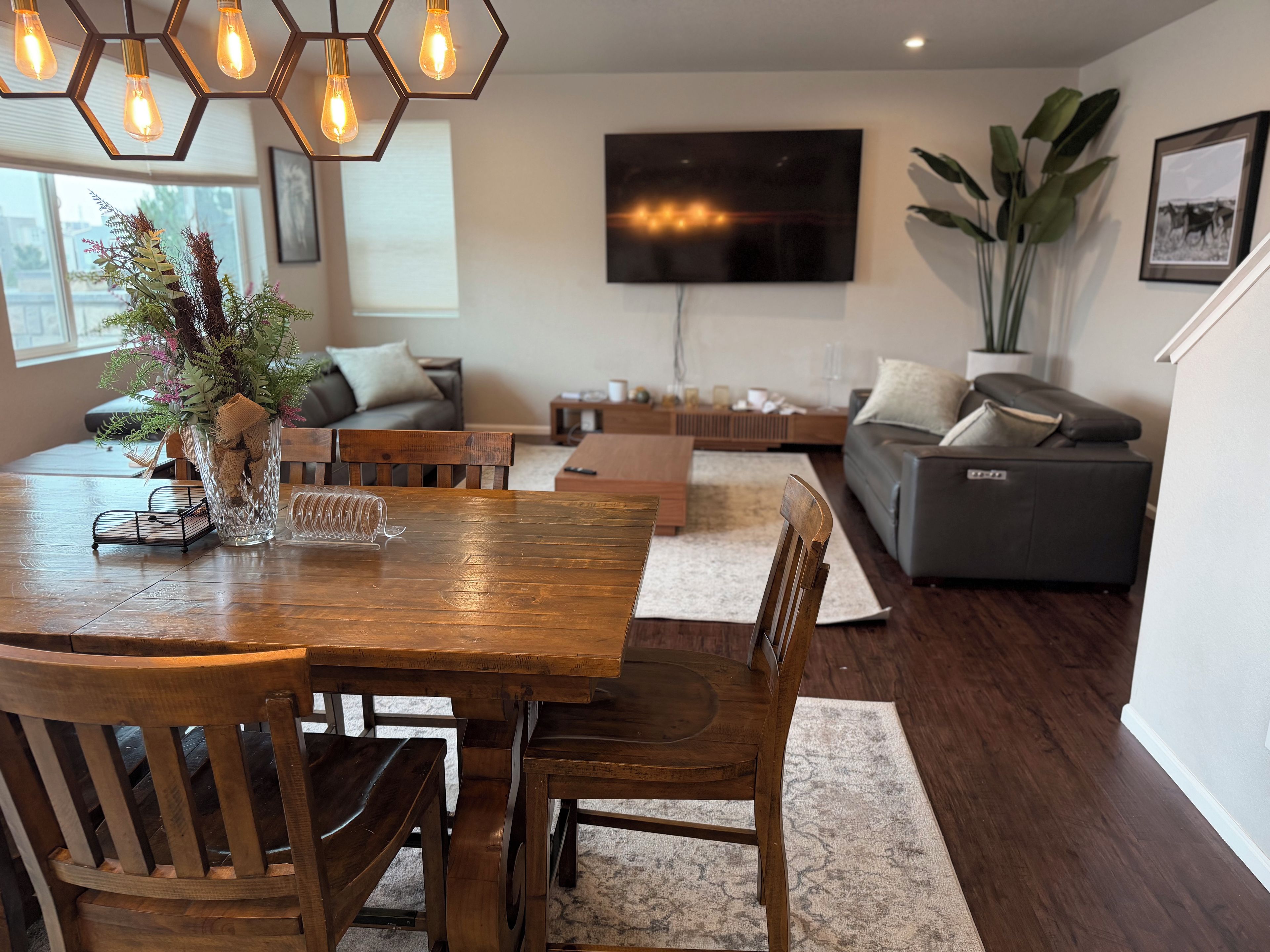 A modern living room with a wooden dining table, chairs, a sofa, and a TV mounted on the wall.