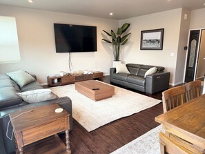 Uma sala de estar moderna com televisão de tela plana, um sofá cinza, uma mesa de centro de madeira e uma mesa de jantar com cadeiras.