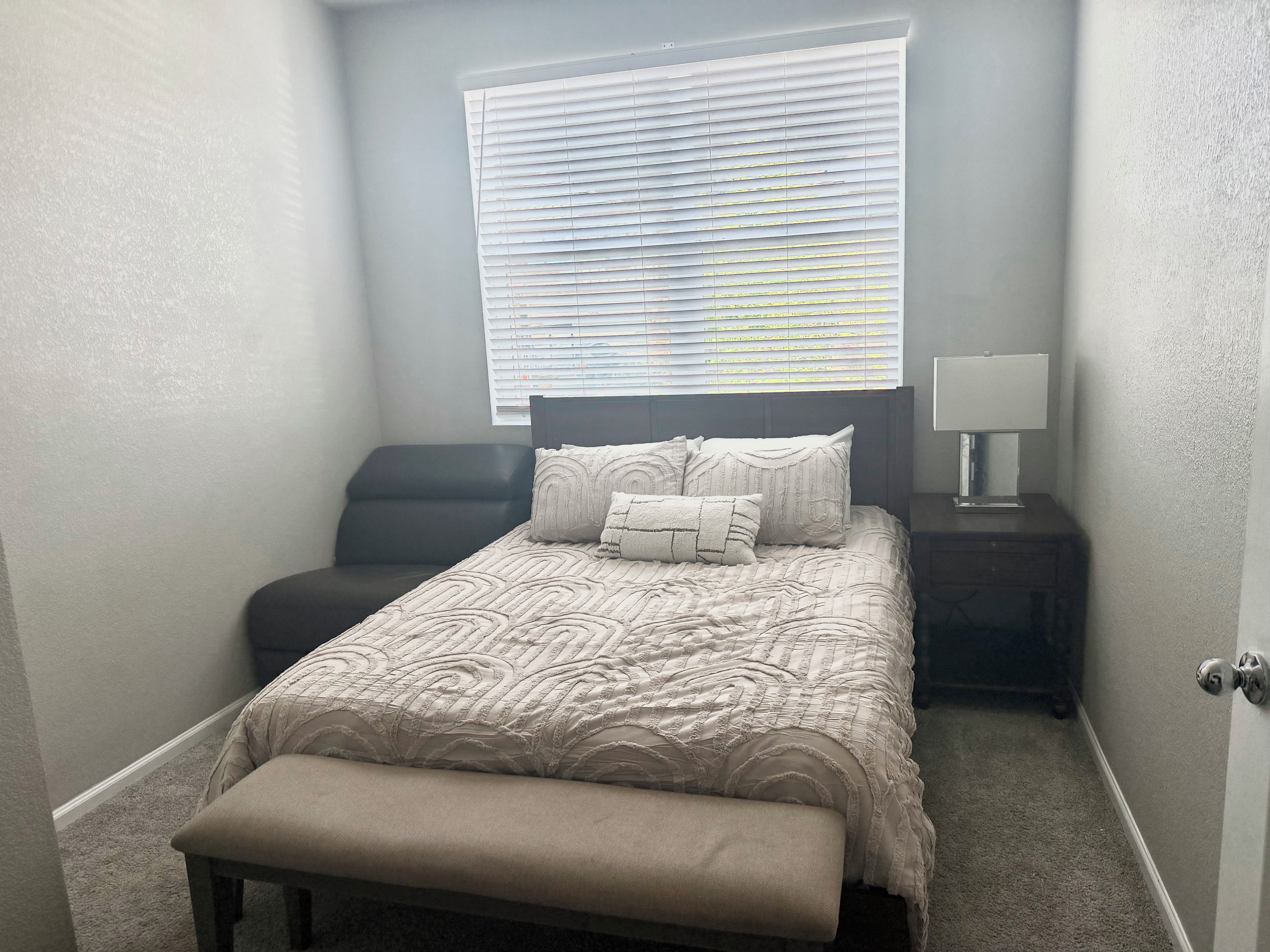 Un dormitorio con cama, un banco, una mesita de noche, una lámpara y una ventana con persianas.