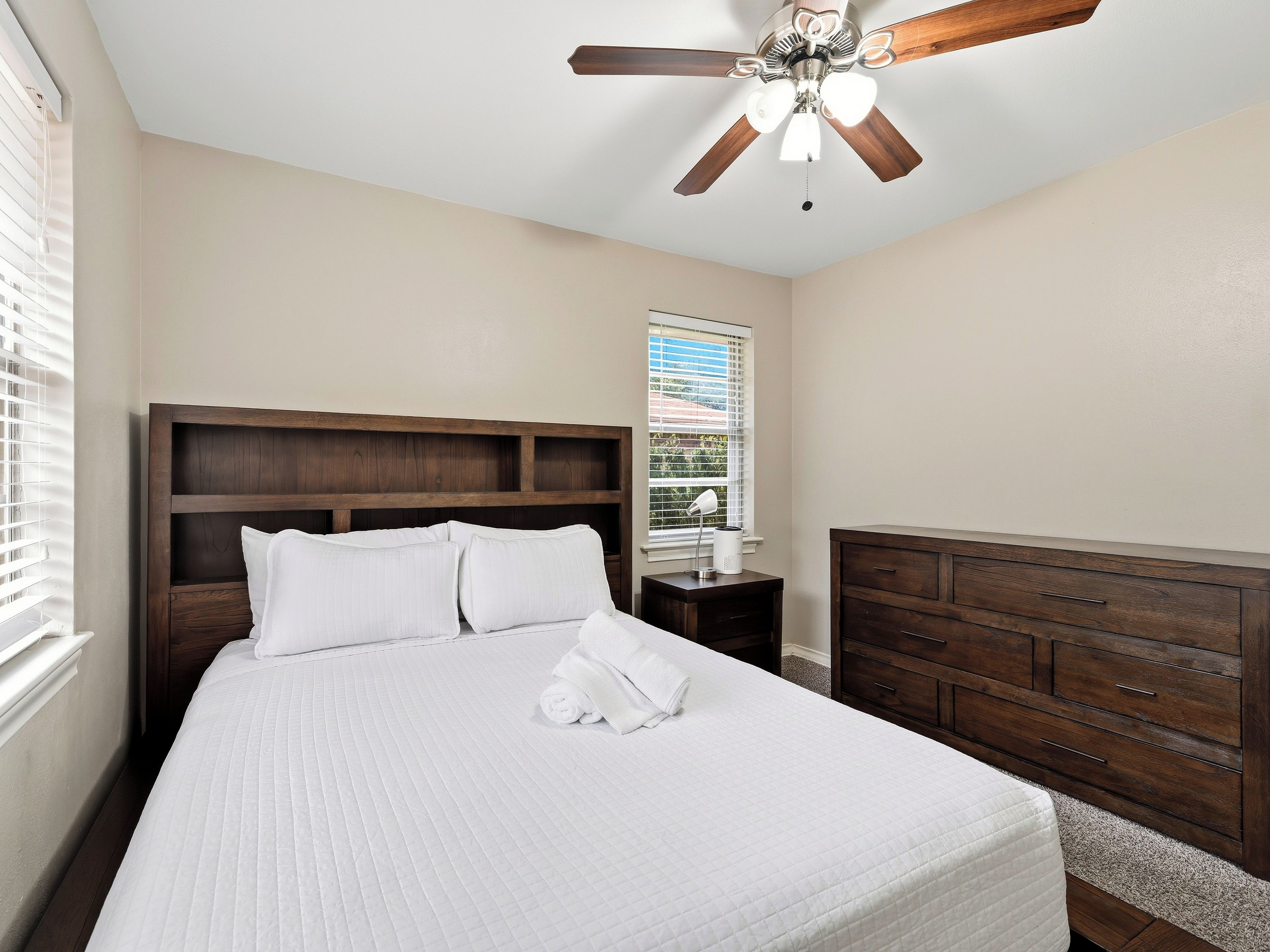 A bedroom with a large bed, a wooden headboard, a ceiling fan, a nightstand, a dresser, and a window with blinds.