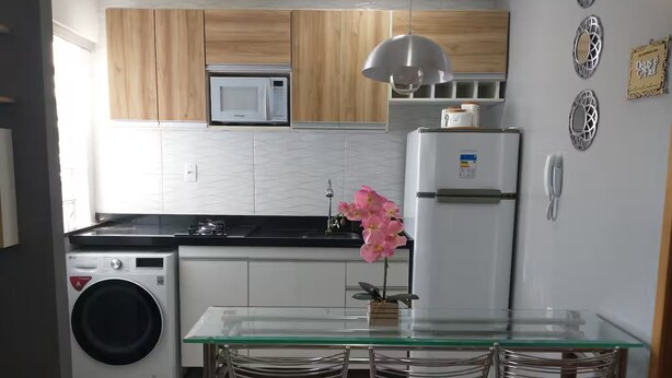 Cozinha compacta com máquina de lavar, micro-ondas, frigorífico e mesa de vidro com cadeiras.
