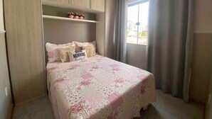 Quarto com cama, armário embutido, prateleira com objetos decorativos e janela com cortinas.