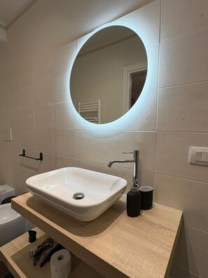 A bathroom with a white sink, a round mirror with a light, a towel rack, and a toilet. - Diamante | Urban Suite (Valenza)