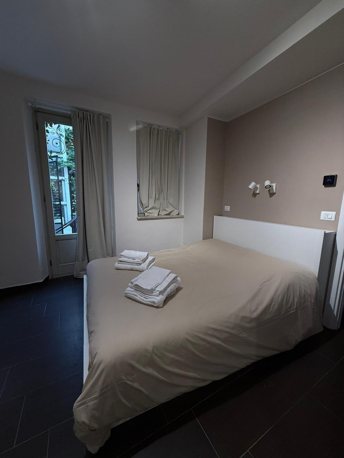 A hotel room with a large bed, a window with curtains, a wall-mounted thermostat, and a door leading outside.