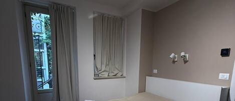 A hotel room with a large bed, a window with curtains, a wall-mounted thermostat, and a door leading outside.