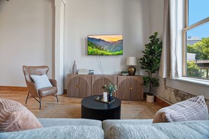 Olohuone, jossa on sohva, nojatuoli, sohvapöytä, kaappi, televisio ja ikkuna, josta on näkymä vehreään maisemaan.