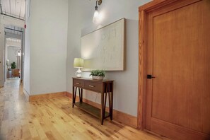 A hallway with a wooden floor, a wooden door, a wall-mounted lamp, and a small table with a lamp and a plant. - Historic Loft, Sleeps 4, Near Downtown Knoxville (Knoxville)