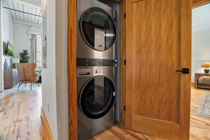 A laundry room with two front-loading washing machines, a wooden door, and a hallway leading to a living area with a chair and a lamp. - Historic Loft, Sleeps 4, Near Downtown Knoxville (Knoxville)