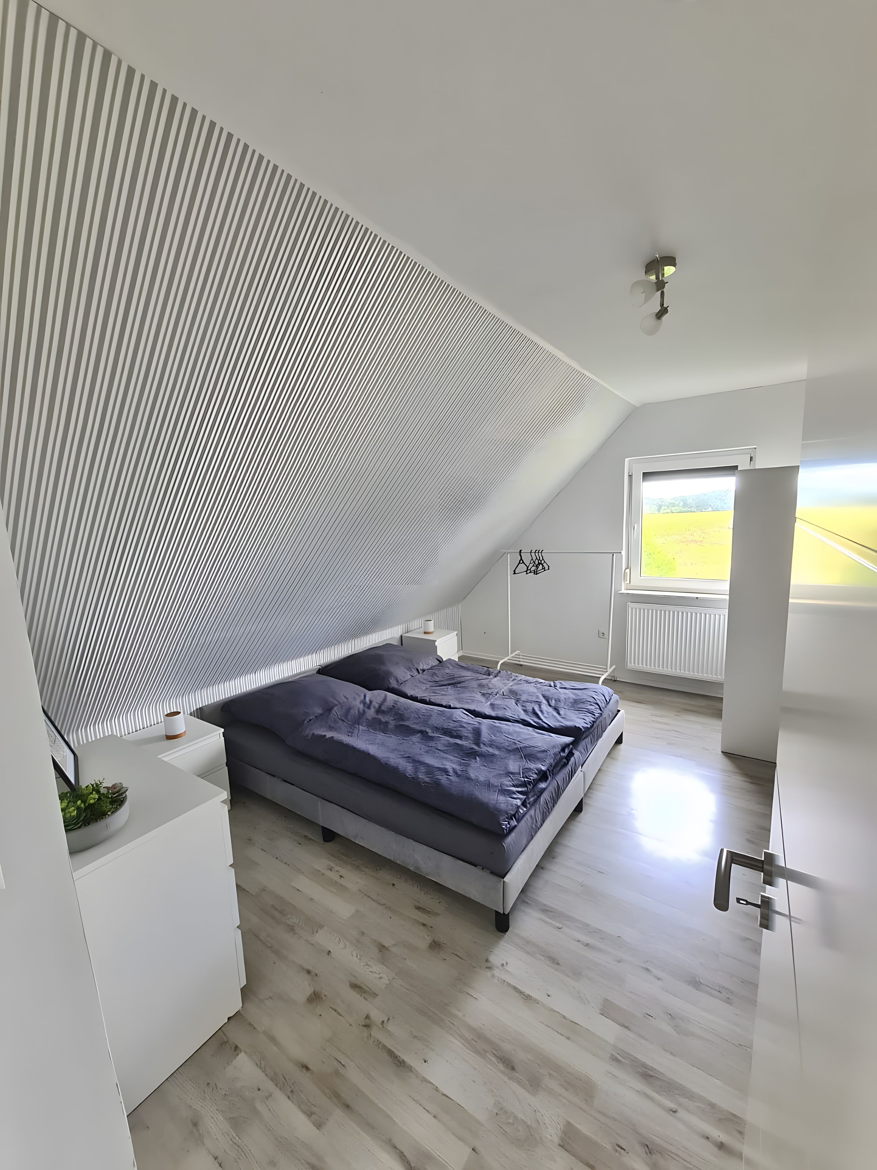 Ein modernes Schlafzimmer mit schrägen Wänden, einem Bett, einem Nachttisch und einem Fenster mit Blick ins Freie.